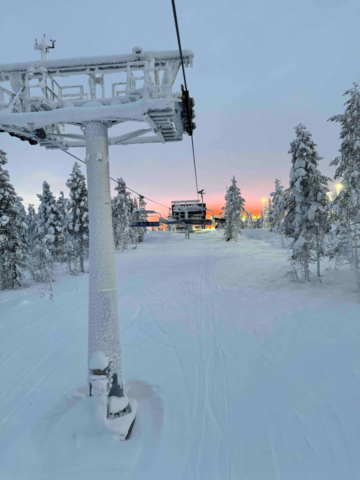 A frosted chairlift tower over a groomed slope, with orange light low behind the trees